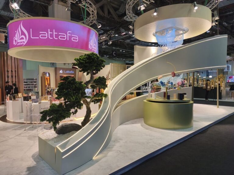 A white sculptural exhibition stand with a curved staircase, pink overhead sign and gold display counter.