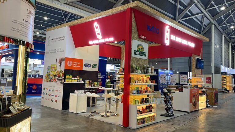 A red corner exhibition pavilion with illuminated signage, retail product shelves and white meeting tables.